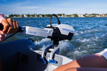 Lähikuva sähköperämoottorista kiinni veneen perässä. Taustalla näkyy peräaallot ja rantakaupunkia.