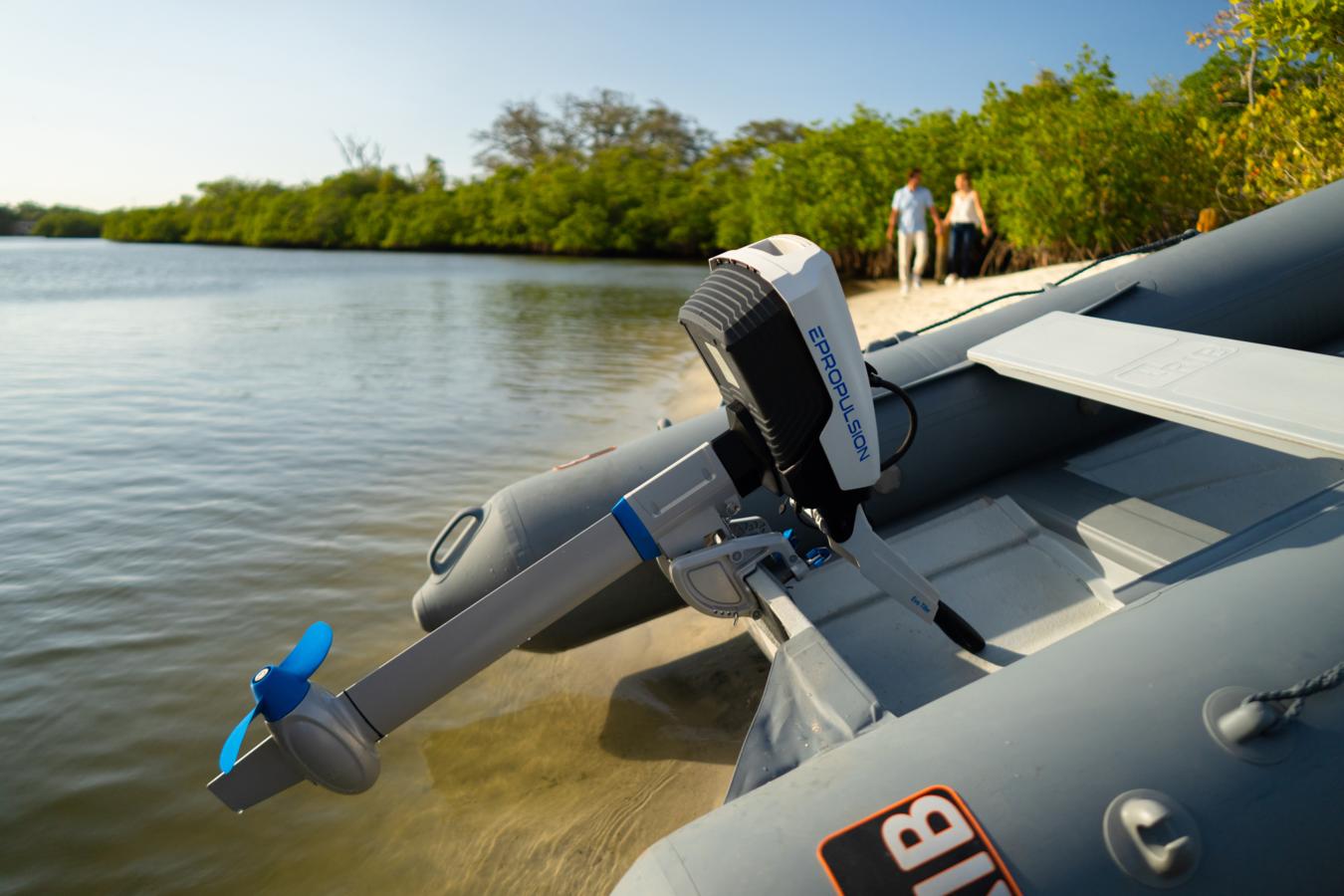 Lähikuva rannalle vedetystä kumiveneen perästä, jossa kiinni sähköperämoottori. Taustalla pariskunta kävelee käsikädessä.