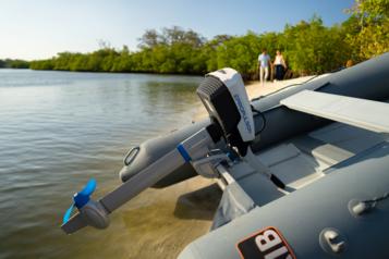 Lähikuva rannalle vedetystä kumiveneen perästä, jossa kiinni sähköperämoottori. Taustalla pariskunta kävelee käsikädessä.