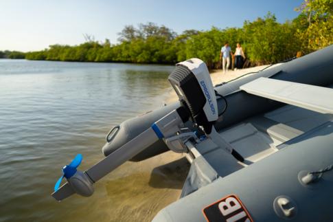 Lähikuva rannalle vedetystä kumiveneen perästä, jossa kiinni sähköperämoottori. Taustalla pariskunta kävelee käsikädessä.