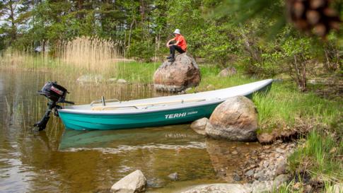 The Terhi Saiman Verde rowing boat at the shore. A teenage boy is sitting relaxed on a large rock behind.